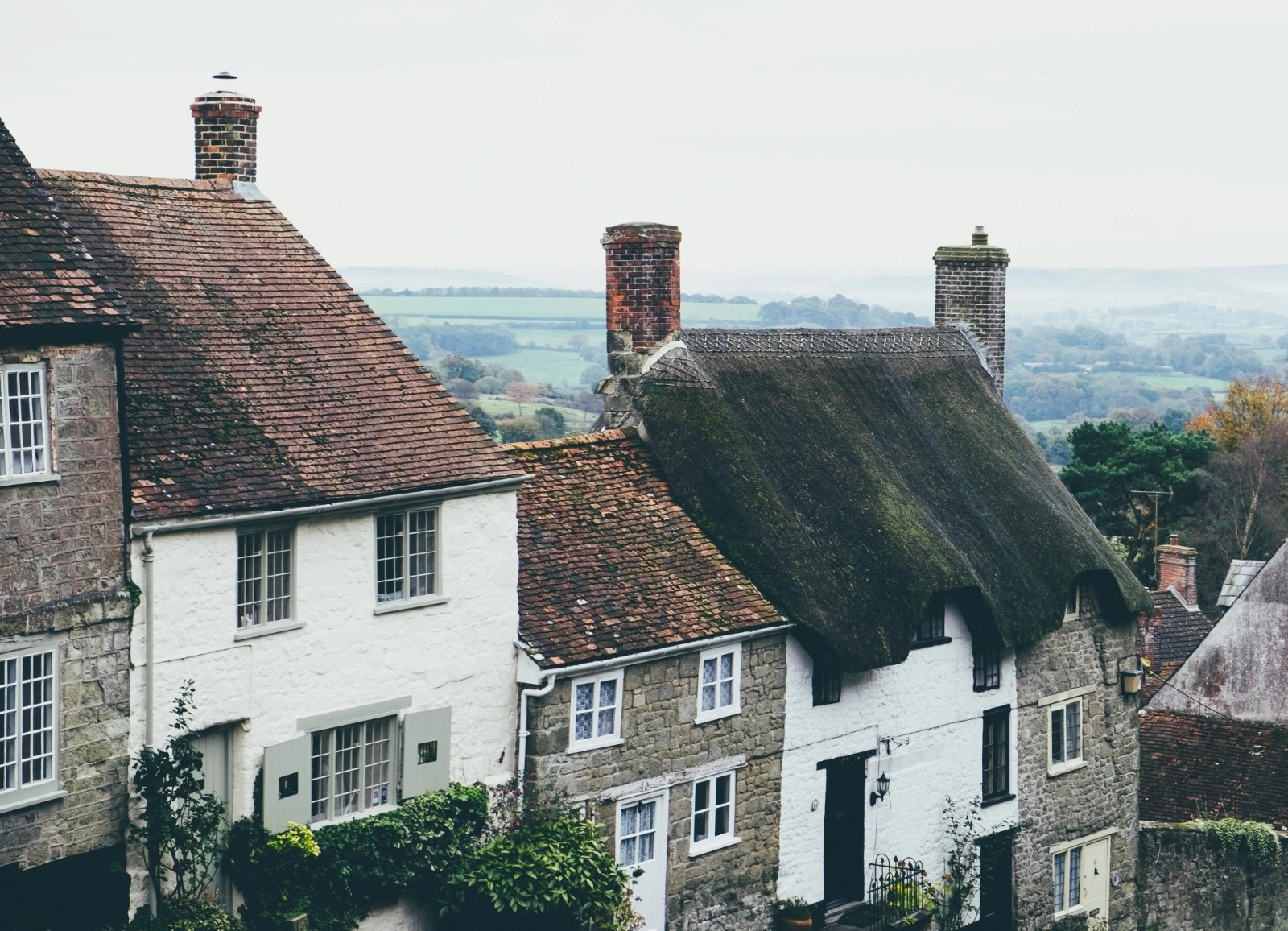 Row of old stone and whitewashed cottages with clay tile and thatched roofs, looking out over misty green countryside