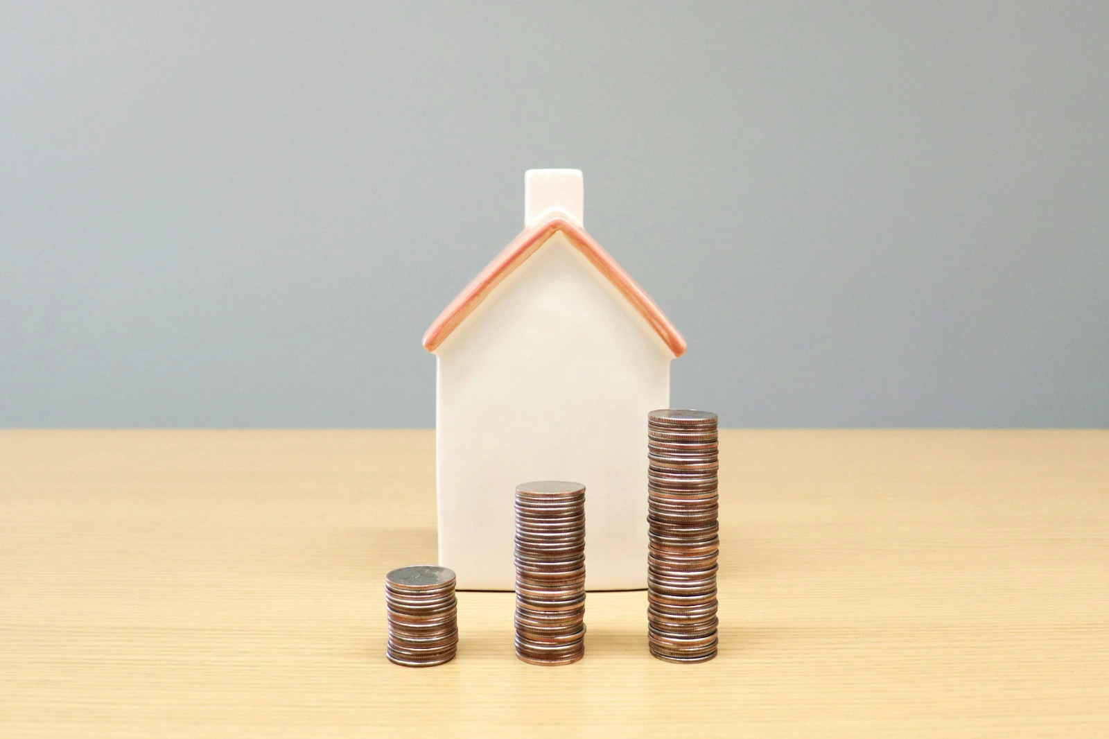 Small ceramic model house on a wooden table with three stacks of coins rising in height beside it